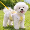 A fluffy white dog wearing a pink lace ruffle plaid harness vest with an angel heart design outdoors.