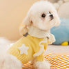 A fluffy white dog wearing a yellow knit sweater with white star accents, sitting on an orange plaid blanket.