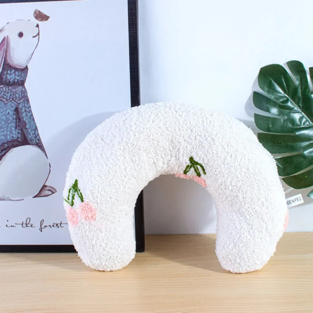 A white U-shaped cat sleeping pillow with pink floral embroidery rests on a wooden surface next to a framed illustration.