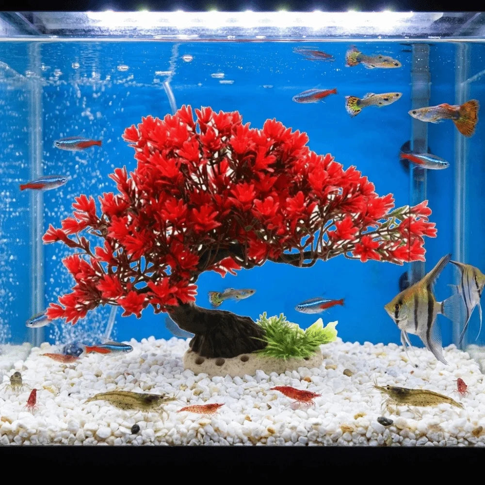 A vibrant red artificial pine tree plant is displayed in a clear aquarium setup with small fish and white gravel.