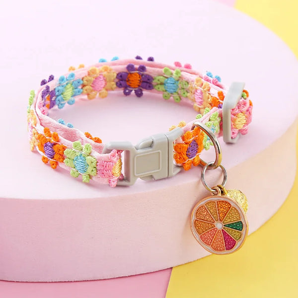 A colorful crocheted pet collar with a bell attached, displayed on a pink surface.