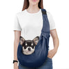A woman wearing a white t-shirt and jeans is holding a navy blue dog sling carrier with a small dog attached.