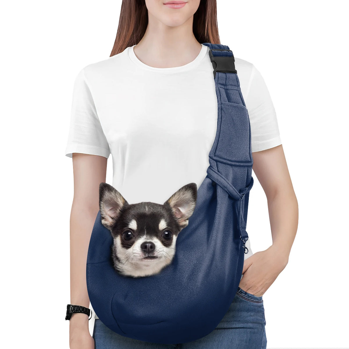 A woman wearing a white t-shirt and jeans is holding a navy blue dog sling carrier with a small dog attached.