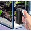 A hand holds a black magnetic aquarium cleaning brush next to a glass tank with rocks and aquatic plants.