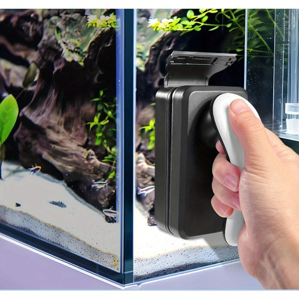 A hand holds a black magnetic aquarium cleaning brush next to a glass tank with rocks and aquatic plants.