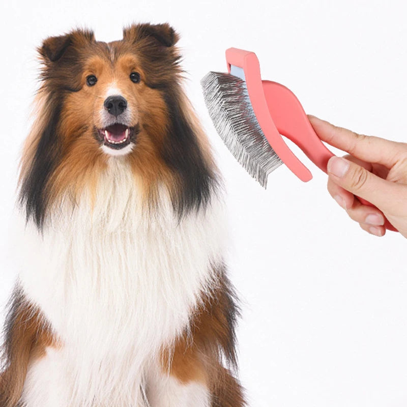 A hand holds a pink grooming rake with stainless steel teeth next to a fluffy brown and white dog.