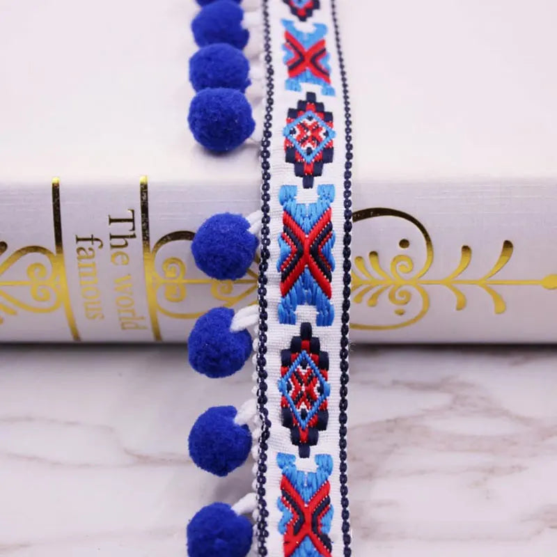 Close-up of a blue and red geometric pom pom fringe lace trim with gold thread on a white fabric roll.