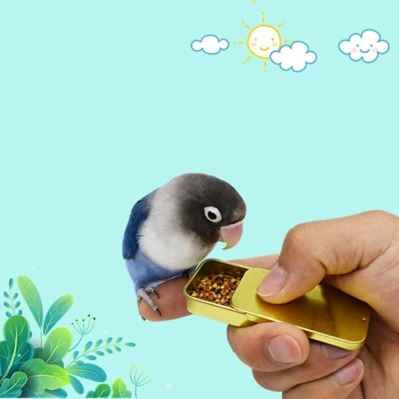 A hand holds a gold-colored treat dispenser with a small parrot sitting next to it against a light blue background.