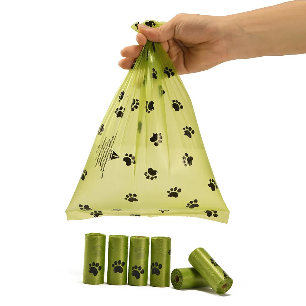 A hand is holding a clear, biodegradable dog poop bag with black paw prints on it, next to several rolls of the same bags.