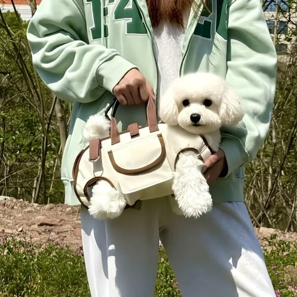 A person holds a beige pet sling carrier for a small white dog against a blurred outdoor background.