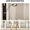 A light tan dog stands inside a white indoor pet playpen with vertical slats on a wooden floor.