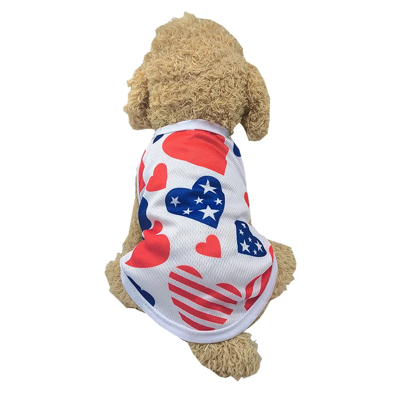 A light brown dog wearing a patriotic Independence Day vest featuring red, white, and blue hearts and stars.