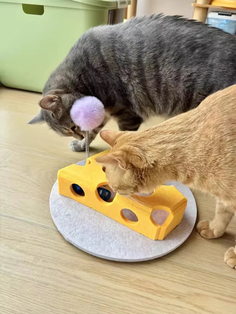 A gray cat is interacting with a yellow felt maze play box featuring interactive balls and a purple teaser toy.