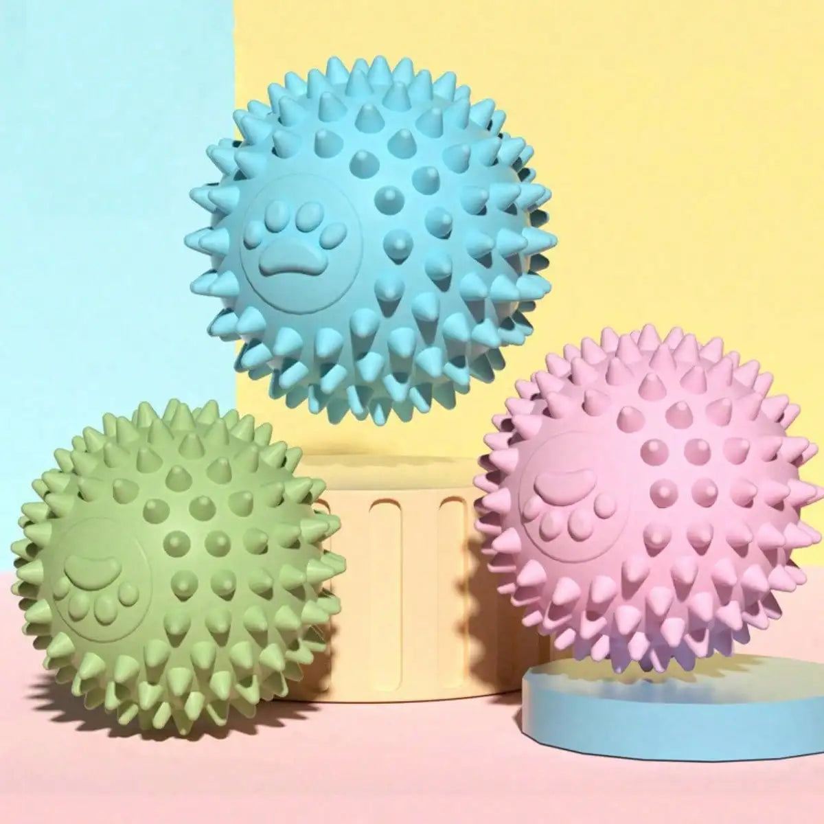Three rubber balls for dogs in blue, pink, and green are displayed against a pastel background.