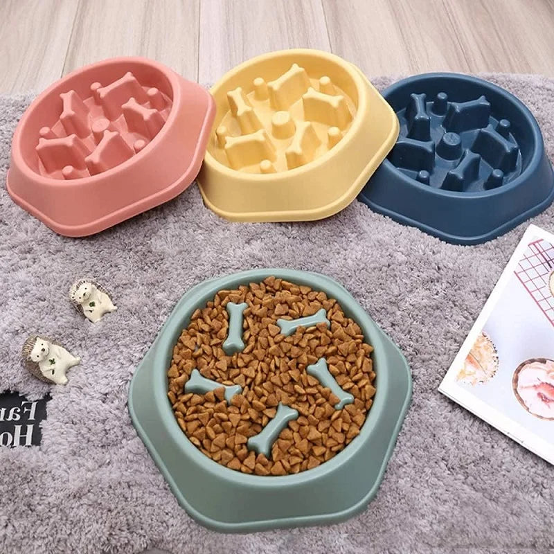 A set of three plastic dog bowls in pink, yellow, and blue, along with a brown slow feeder bowl on a gray fabric.