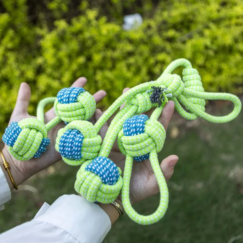 A hand holds a bright lime green cotton rope dog toy with blue accents, set against a blurred green outdoor background.