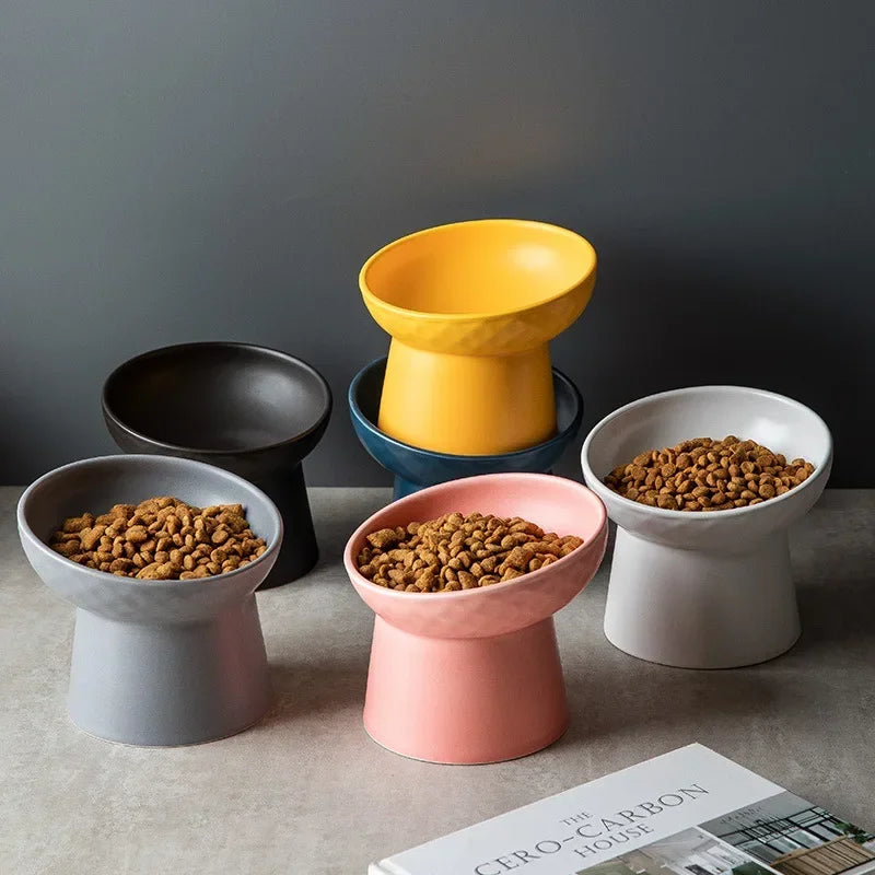 A collection of elevated ceramic food bowls in yellow, black, and pink sitting on a neutral surface.