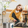 A stainless steel pet water fountain is sitting on a wooden table next to a sleeping cat.