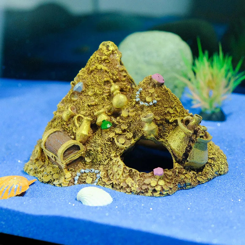 A miniature mountain cave decoration featuring a treasure chest design, resting on blue fabric with a seashell nearby.
