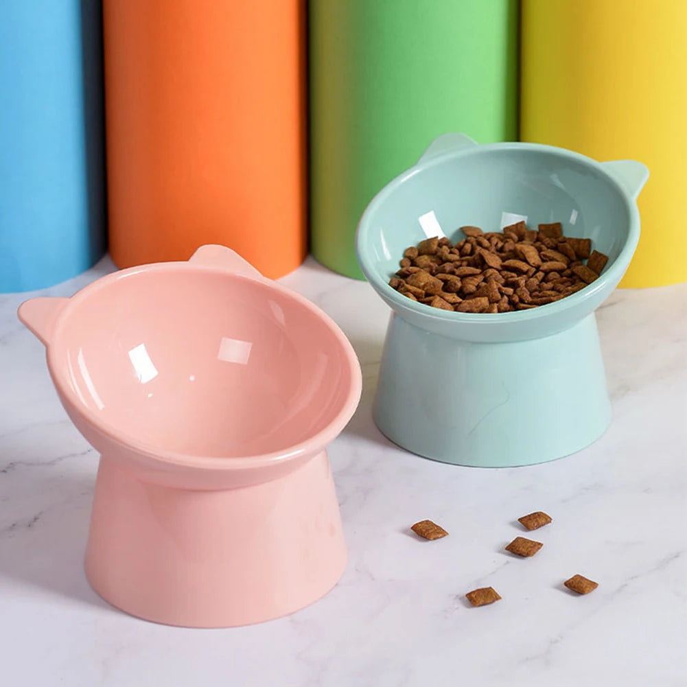 A pink ergonomic tilted cat food bowl sits on a white marble surface next to a green bowl filled with dry cat food.