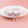 Pink lace bow cat collar with a silver bead charm and dangling pearl bells displayed on a white plate.