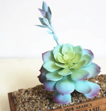 A light blue artificial succulent plant with fleshy leaves is displayed on a bed of small pebbles.
