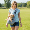 A woman is holding a light blue pet carrier bag with a safety harness and a golden-colored puppy outdoors in a grassy field.