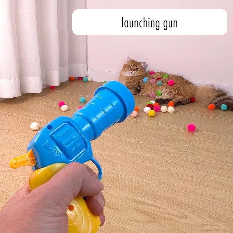 A hand is holding a blue launching gun, aiming it at colorful balls near a cat.