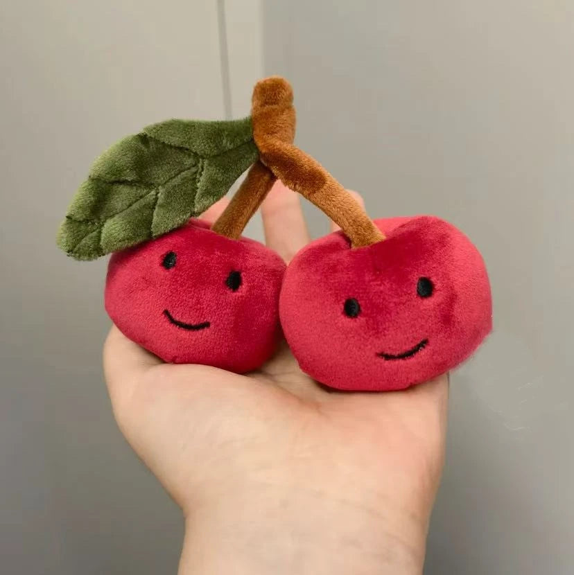 A pair of adorable red cherry plush keychains with green leaves and brown stems held in a hand.