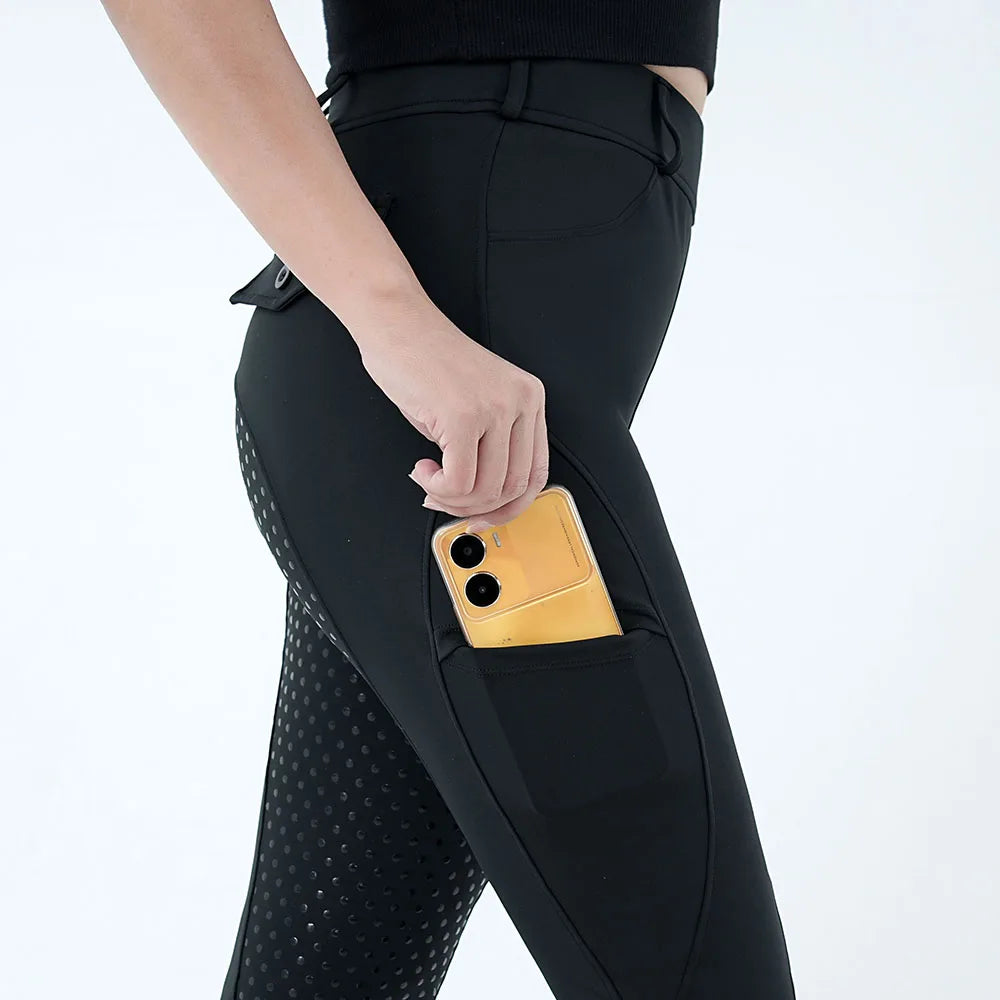 A close-up of a woman's hand inserting a gold smartphone into a pocket of black riding breeches.