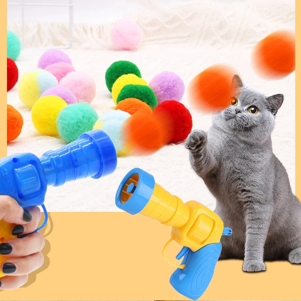 A gray cat is interacting with a blue and yellow toy gun, surrounded by colorful soft plush balls.