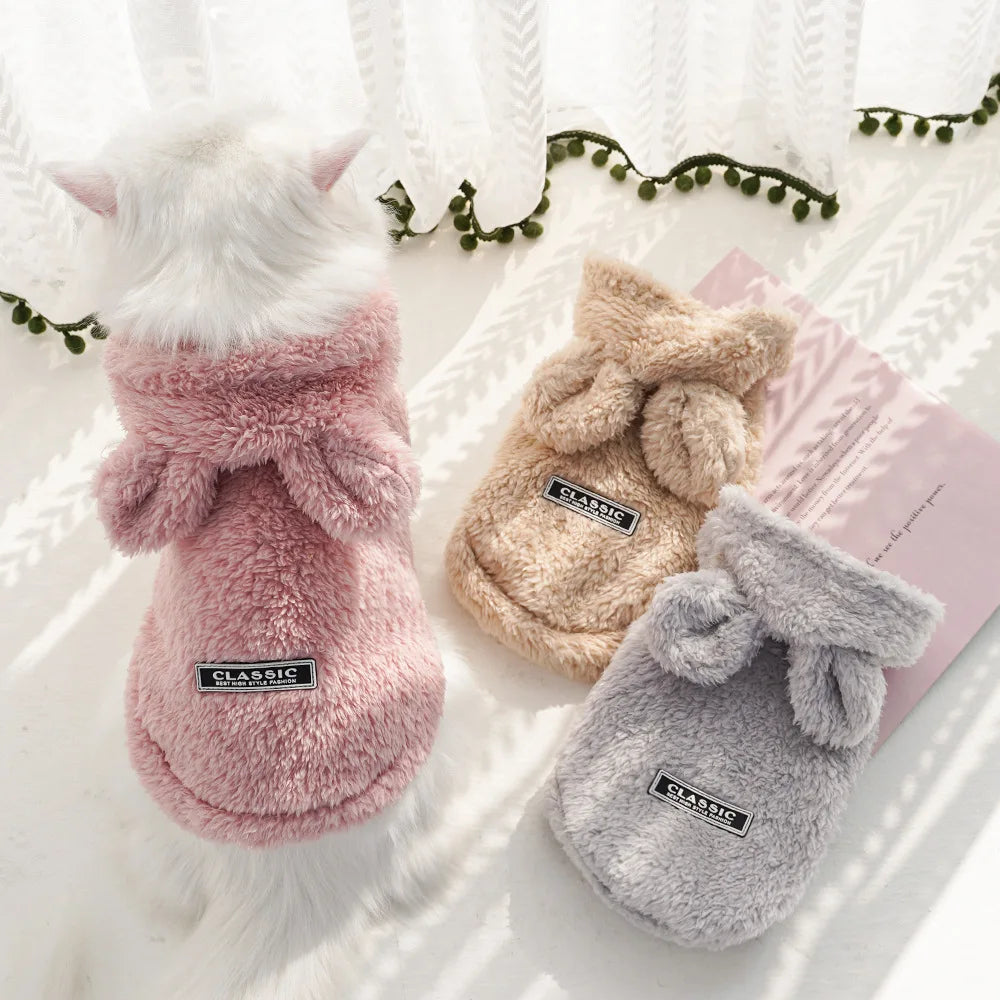 Drei kuschelige Fleece-Sweatshirts für Haustiere in rosa, beige und grau auf einer weißen Oberfläche.
