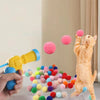 A hand is launching a pink plush ball towards a fluffy orange cat in a light-colored room.