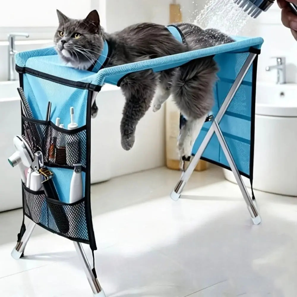 A gray cat is resting in a blue pet grooming hammock suspended from a folding metal support frame.