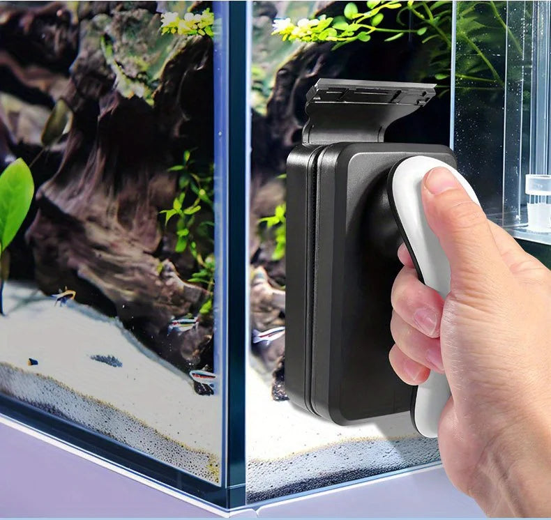 A hand is using a black and white magnetic aquarium cleaning brush to clean a glass tank.
