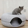 A gray cat is sitting inside a round, soft pet bed with an entrance tunnel.