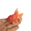 Ein kleines, leuchtendes, orange-rotes, leuchtstoffhaltiges Goldfisch-Dekor wird in der Hand einer Person gehalten.