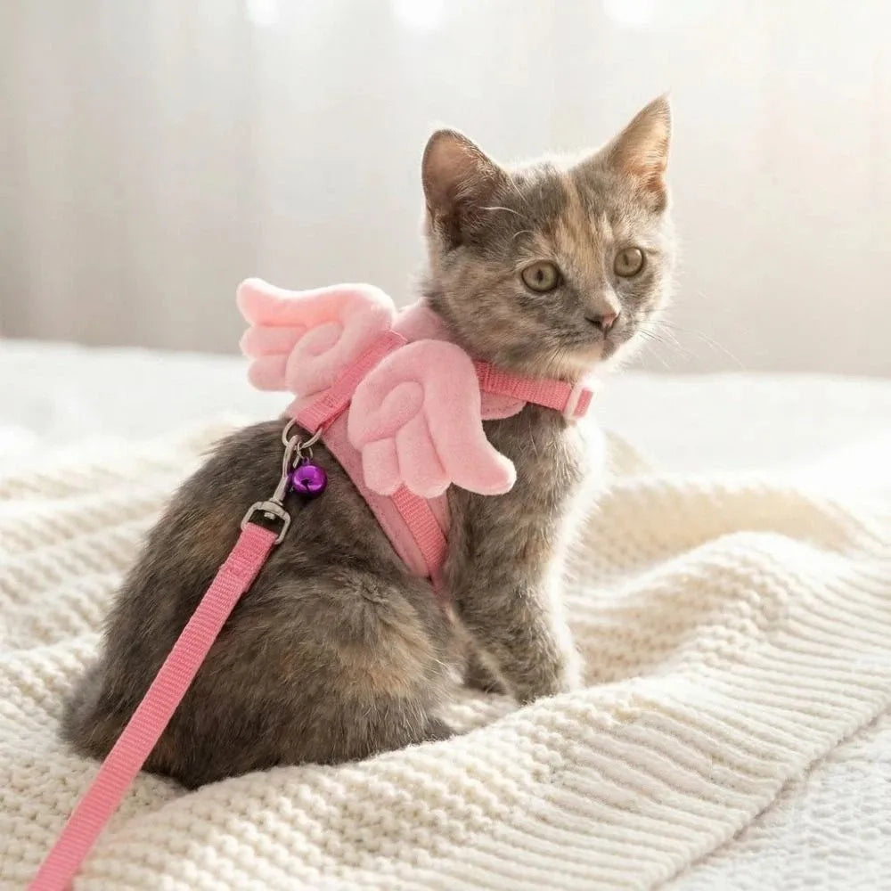 A small gray kitten wearing a pink, cartoon-style pet harness with a leash, lying on a white blanket.