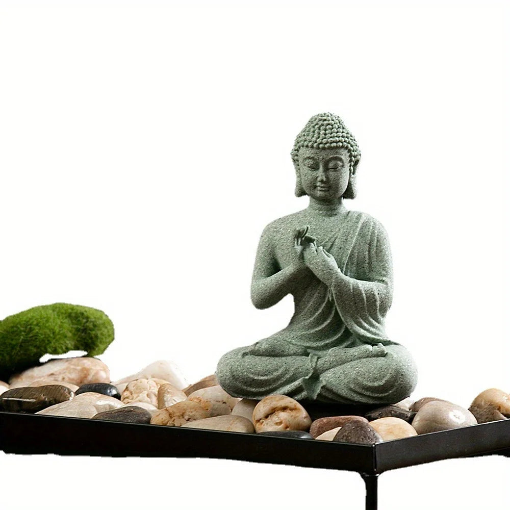 A small, greenish-gray Buddha stone statue seated in meditation on a dark tray surrounded by smooth stones.
