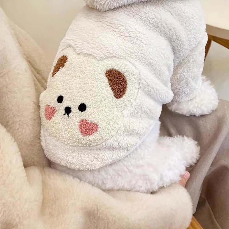 Un pull à capuche pour chien en polaire blanche avec des marques brunes, tenu dans un tissu beige doux.