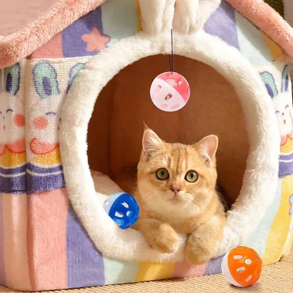 A small orange tabby kitten sits inside a white felt cat house, looking at a pink jingle bell toy hanging in front of it.