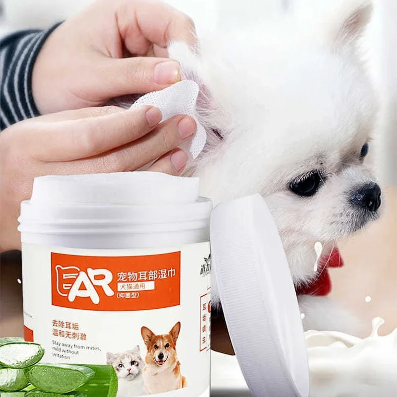A hand is cleaning a white dog's ear with a white wipe, next to a container of pet eye and ear cleaning wipes.