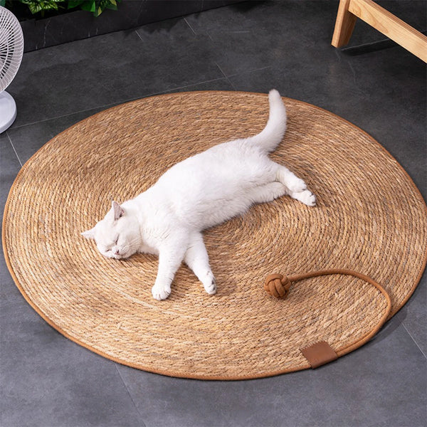 A white cat lying on a round natural rattan scratching mat with a wooden ball nearby.