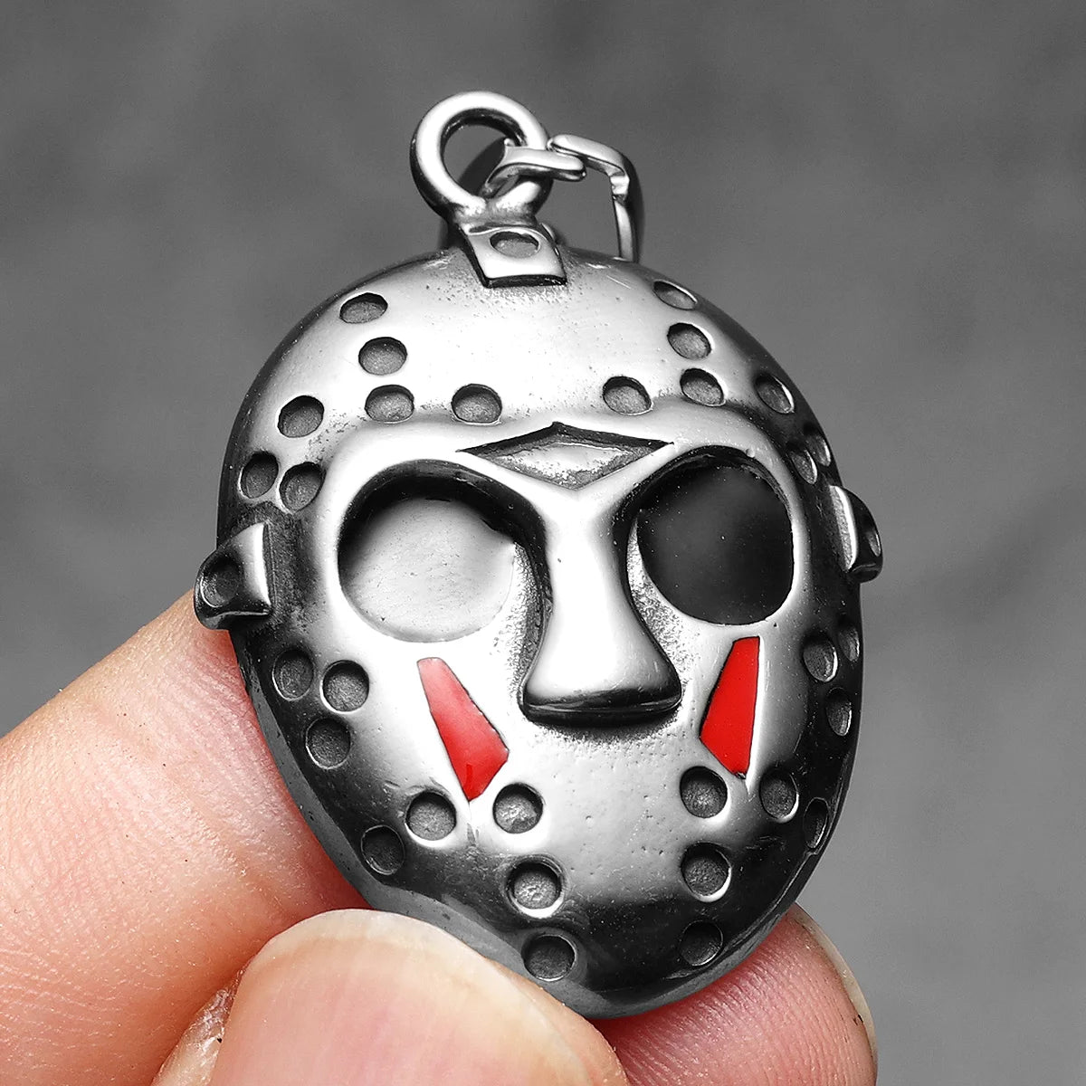 A silver stainless steel pendant necklace shaped like Jason Mask's mask, held by a finger.