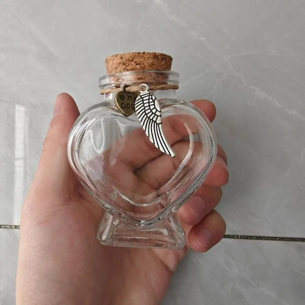 A hand holds a clear glass heart-shaped jar with a cork stopper and a hanging charm featuring a feather.