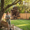 A woman is sitting on a wooden bench in a garden with a rustic wooden bird house hanging from a tree branch.