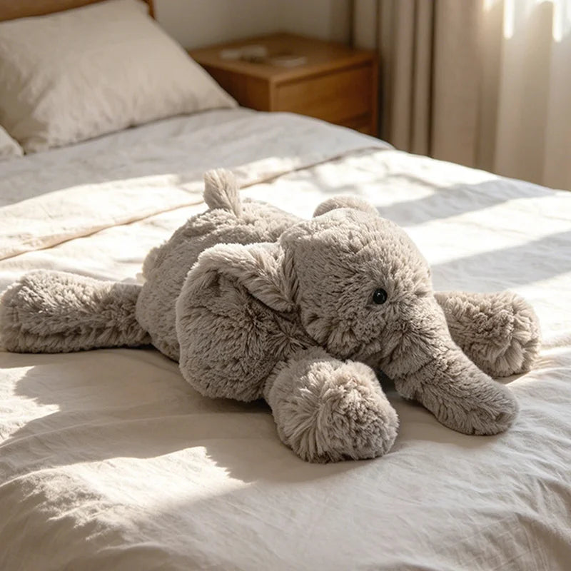 A light gray elephant plush toy lying on a white bed sheet.