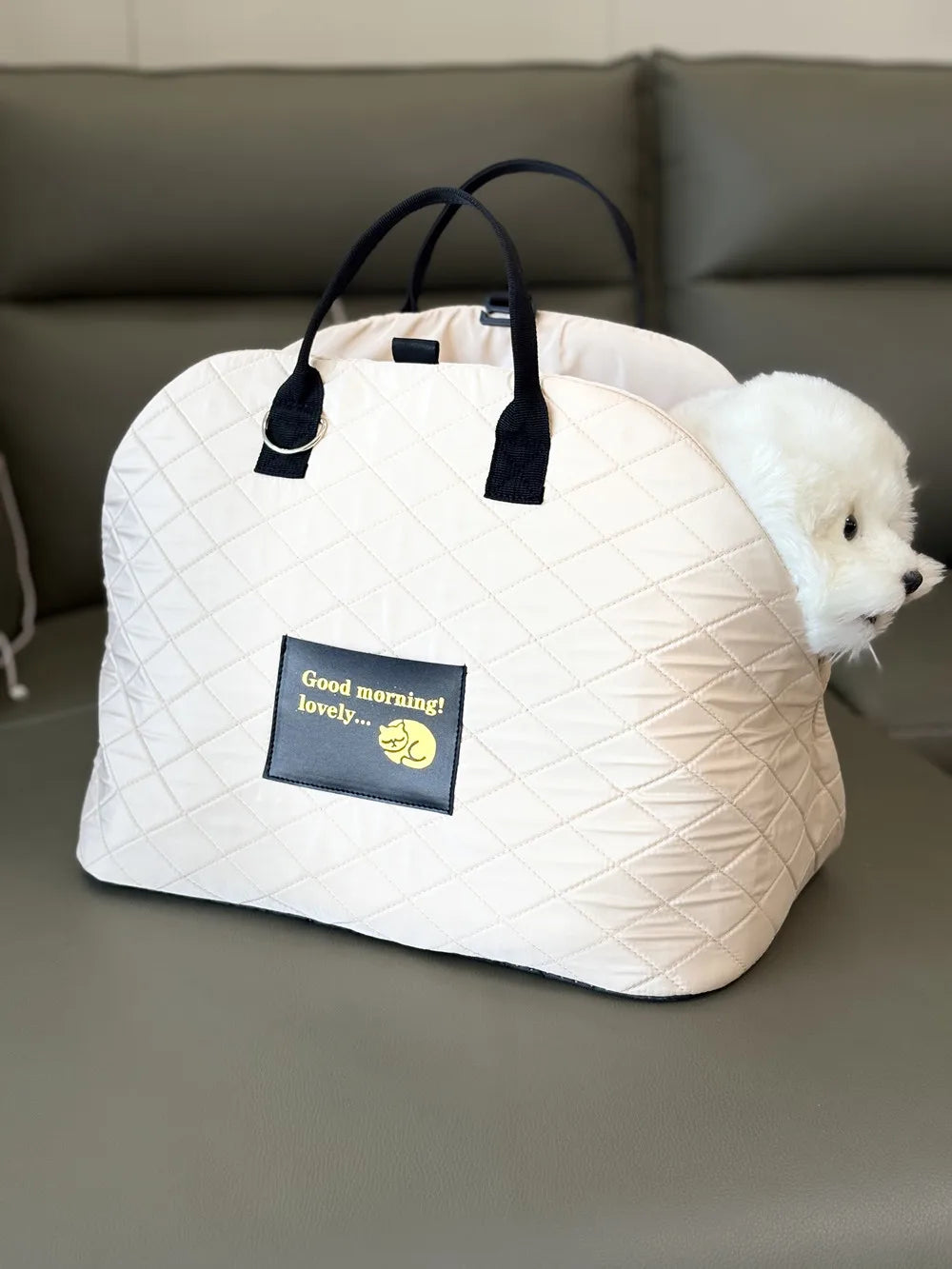 A cream quilted soft-sided pet carrier bag with black handles resting on a gray couch next to a white dog.