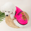 A white fluffy dog wearing a pink pet vest with a brown bear print on a woven mat.
