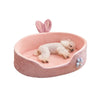 A pink plush pet bed with a removable cover and pink bunny ears, featuring a fluffy white dog lying inside.